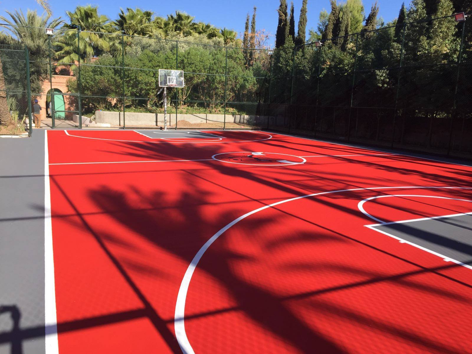 Red outdoor basketball court with palm shadows.